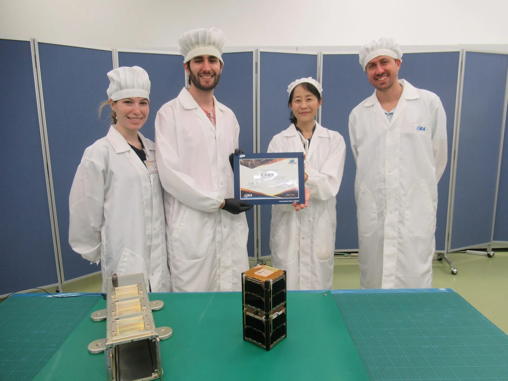 Four people in cleanroom coats holding the JAXA handover certificate for CoRAL, with the flight model and its deployer on the bench in front of them